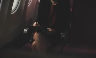 Blonde female tourist checking incoming notification on smartphone sitting on seat of airplane with netbook.Young businesswoman share media from telephone on laptop computer during plane flight photo