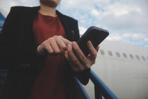 joven asiático mujer en internacional aeropuerto, utilizando móvil teléfono inteligente y comprobación vuelo a el vuelo información tablero foto