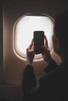 Blonde female tourist checking incoming notification on smartphone sitting on seat of airplane with netbook.Young businesswoman share media from telephone on laptop computer during plane flight photo