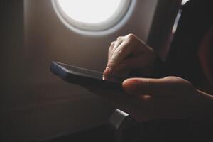 Blonde female tourist checking incoming notification on smartphone sitting on seat of airplane with netbook.Young businesswoman share media from telephone on laptop computer during plane flight photo