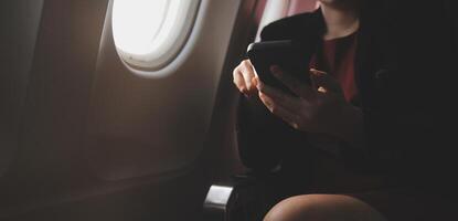Blonde female tourist checking incoming notification on smartphone sitting on seat of airplane with netbook.Young businesswoman share media from telephone on laptop computer during plane flight photo