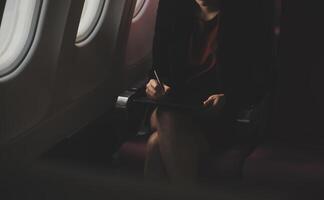 Blonde female tourist checking incoming notification on smartphone sitting on seat of airplane with netbook.Young businesswoman share media from telephone on laptop computer during plane flight photo