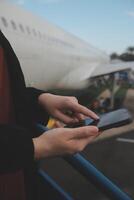 joven asiático mujer en internacional aeropuerto, utilizando móvil teléfono inteligente y comprobación vuelo a el vuelo información tablero foto