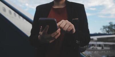 joven asiático mujer en internacional aeropuerto, utilizando móvil teléfono inteligente y comprobación vuelo a el vuelo información tablero foto