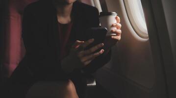 Blonde female tourist checking incoming notification on smartphone sitting on seat of airplane with netbook.Young businesswoman share media from telephone on laptop computer during plane flight photo