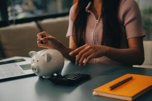 joven asiático mujer ahorro para Jubilación ahorro dinero mediante un cerdito banco y tomando notas en computadora portátil, ahorros concepto. foto