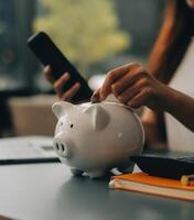 Young Asian woman saving for retirement Saving money through a piggy bank and taking notes on notebook, savings concept. photo