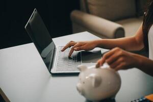 joven asiático mujer ahorro para Jubilación ahorro dinero mediante un cerdito banco y tomando notas en computadora portátil, ahorros concepto. foto