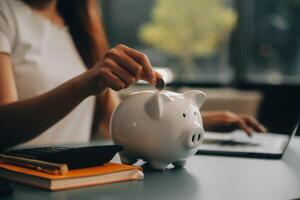 joven asiático mujer ahorro para Jubilación ahorro dinero mediante un cerdito banco y tomando notas en computadora portátil, ahorros concepto. foto