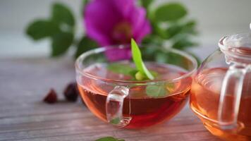 gebrouwen rozenbottel thee in een glas theepot met rozenbottel bloemen en munt, Aan een houten tafel. video