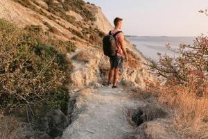 Sporty man in a T-shirt and shorts with backpack hiking on the coastline. photo