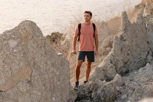 Man in shorts with backpack staying on rocky coastline with warm sun light. photo