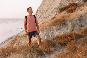 deportivo hombre en un camiseta y pantalones cortos con mochila quedarse en el línea costera a calentar puesta de sol. foto