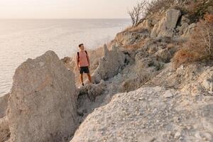 Man in a T-shirt and shorts with backpack staying on rocky coastline with warm sun light. photo