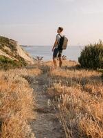 hombre en un camiseta y pantalones cortos con mochila en el línea costera con calentar puesta de sol. foto