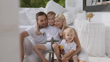 contento joven familia de cuatro con pequeño hermosa niños teniendo divertido mientras sentado en el porche a hogar. video