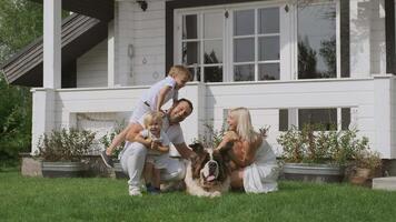 A young family of four in bright clothes with children and a dog having fun sitting on the grass near their white house video