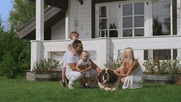 A young family of four with children and a dog having fun sitting on the grass near their white house video