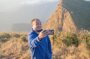 Asian fat man take a selfie with beautiful sunrise of khao San nok wua mountain kanchanaburi.Khao San Nok Wua is the highest mountain in Khao Laem National Park. It is 1767 meters above sea level. photo