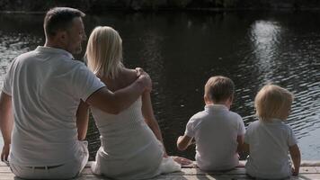 Happy young family mom dad and two children are sitting on the pier near the river. Back view video