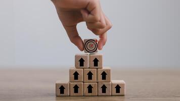 Hand arranges a wooden block with a dartboard icon stacked in a pyramid shape. Goal Achievement and Purposefulness, the challenge in business concept., photo