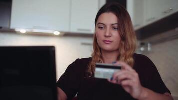 skön ung kvinna med lång mörk hår uppkopplad bank använder sig av en mobil dator på de internet med en kreditera kort på Hem på de tabell har en Bra humör och leende. hd video