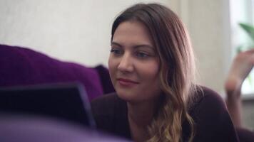 Close-up. A beautiful young girl with long hair lies on the couch and enjoys a tablet moving her legs having a good mood and a smile. HD. video