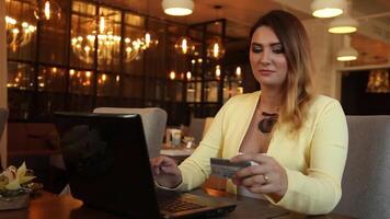 Beautiful business woman in a bright jacket with long blond hair makes an online purchase using a credit card while sitting at a table in a cafe having a good mood. HD. video