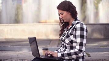 Young attractive girl with long hair works on a mobile computer with a serious look sitting on the porch in the city and holds coffee in her hand. video