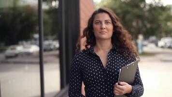 Close-up. A beautiful business woman with loose long hair and a dark suit walks next to an office building outdoors in sunny weather during working hours with documents in hands video