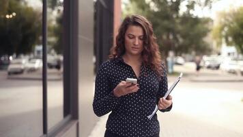 Portrait. Young business woman in a dark suit is walking through the city next to the office with documents and talking on the phone video