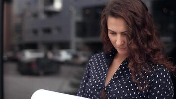 Close-up. Young business woman with long hair and dark shirt looks at documents with a serious look standing next to office building video