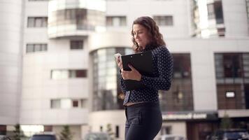 Young attractive business woman in dark clothes and loose hair passes through the city with documents during the day in office hours talking on the phone in a good mood. video