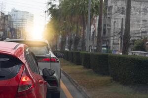 Traffic jam on the road in the city at sunset. Close up rear side of red car with turn on brake light. Beside with island of green grass and trees. And city for background. photo