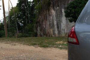 Beside of grey car can see tail light. Parking in the park. Background of dirt roads and high mountains and forests. photo