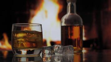 Glass and bottle of whiskey, scotch or bourbon stands on a table against background of a fireplace with a flame. Ice falls into a glass of whiskey. Alcoholic drink and home comfort concept. video