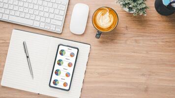 Business analytics concept, Overhead view of a business desk featuring a smartphone displaying pie charts, alongside a keyboard, notebook, coffee cup, and photography equipment. photo