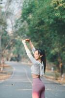 Fitness and running concept, A smiling young woman performs arm stretches on a sunny park path, wearing earphones and sporty attire. photo