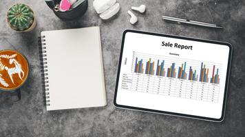 Business analytics concept, Modern tablet displaying a vibrant sales report with bar graphs on a desk with coffee, notebook, and stationery, portraying an efficient work environment. photo