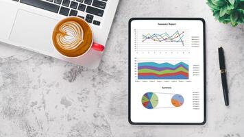 Business analytics concept, A professional workplace featuring a tablet displaying graphs and reports, accompanied by a laptop, a cup of coffee, and a pen on a marble surface. photo