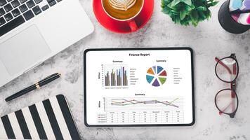 Business analytics concept, A tablet showcasing a finance report with colorful graphs beside a red coffee cup, laptop, and stationery on a marble background. photo