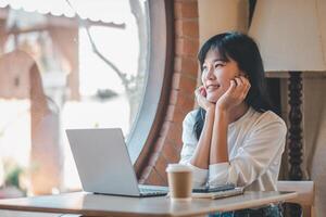 Business freelance concept, A cheerful young woman takes a break at a cozy cafe, smiling while looking out the window with a laptop and coffee on the table. photo