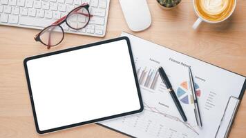 A neatly organized workspace featuring a blank tablet screen, keyboard, glasses, and a detailed finance report, perfect for business analysis and planning. photo