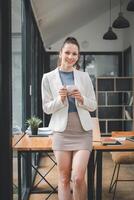 A smiling young professional woman in a modern workspace enjoys a coffee break, embodying confidence and workplace positivity. photo