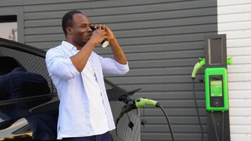 negro hombre hablando en el teléfono y Bebiendo café mientras cargando lujo eléctrico coche a estación video