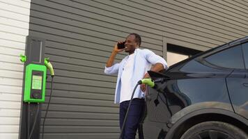 African american man talking on the phone while charging electric car at charging station video