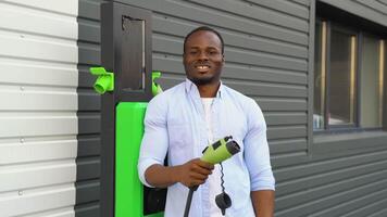 retrato de sonriente negro hombre con un cargador a ev cargando estación video