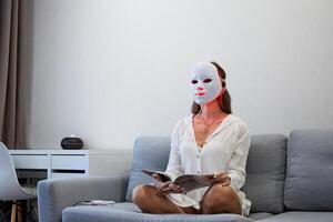A beautiful girl with an LED mask on her head works at home on a laptop. Home skin care concept. photo