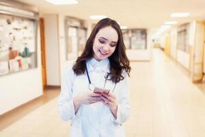 Young female doctor Smiling in a white coat with a phone in hands. Beautiful medical student look at the tablet photo