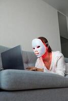 A beautiful girl with an LED mask on her head works at home on a laptop. Home skin care concept. photo